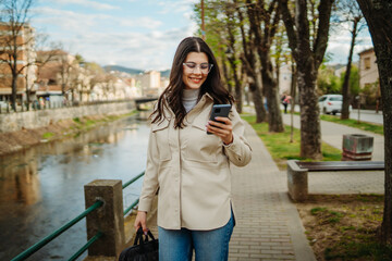 Fototapeta premium Young caucasian woman walk home from work drink coffee and use smartphone 