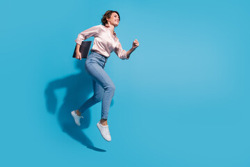 Obraz premium Young woman in casual fashion jumping joyfully against blue background, holding a tablet and wearing stylish outfit