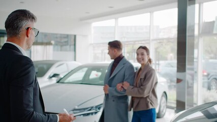 Salesman with clipboard greets young couple in car dealership