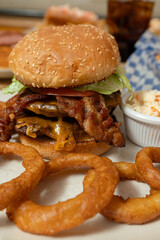 A close-up of a delicious diner-style bacon cheeseburger served on a white plate with crinkle-cut French fries, crispy onion rings, and a side of creamy coleslaw.