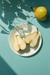 White plate with green pear half fruit on the blue background with leaves shadows.