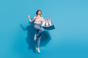 Joyful young woman jumping with shopping bags against blue background wearing stylish satin blouse