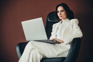 Stunning businesswoman in a white suit working on a laptop while seated on a stylish chair in a...