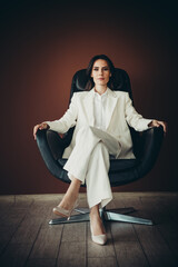 Beautiful young businesswoman in elegant white suit seated in black leather chair against brown...