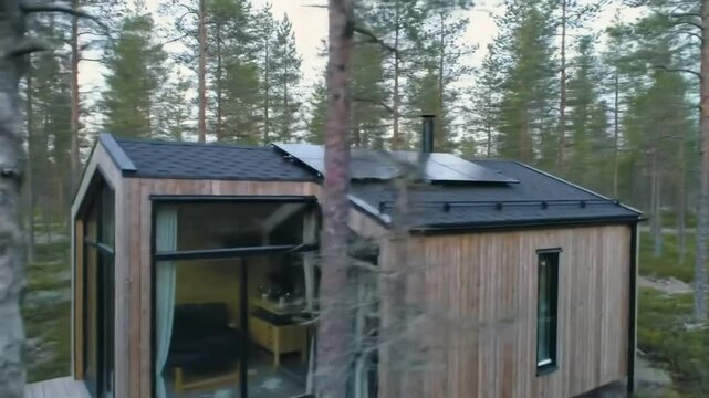 A small modern cabin with solar panels on the roof sits amongst tall thin trees. The building has light wood cladding and large glass windows.

