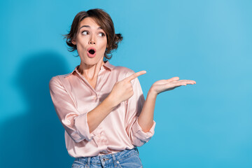 Surprised young woman pointing to copy space on blue background, wearing a satin pink blouse and...