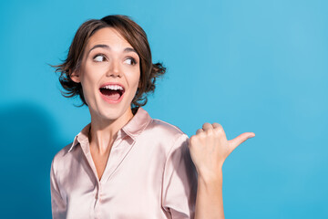 Young woman with stylish bob hairstyle in satin blouse gesturing with thumb and smiling joyfully against vibrant blue backdrop