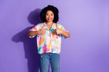 Smiling young woman showing thumbs up gesture against a vivid violet background wearing casual...
