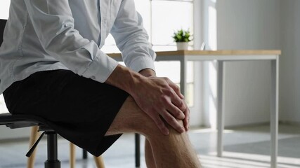 Office professional experiencing persistent knee pain while seated, gently massaging affected area to alleviate discomfort in contemporary workplace environment