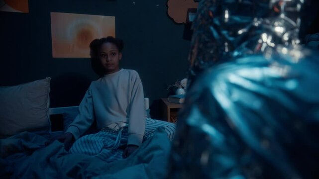 Frightened Black girl in pyjamas sitting on bed in dark room at night trying to hide from unrecognizable monster approaching her with creepy claws under flickering light