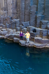 Two explorers stand hand in hand on the rocky banks of Studlagil Canyon, surrounded by striking...