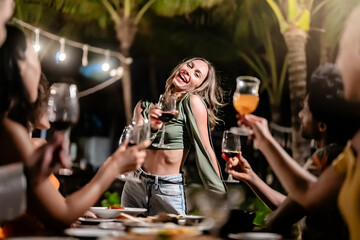 Group of diverse friends raising glasses in a joyful toast at a tropical outdoor party, celebrating together under palm trees and string lights