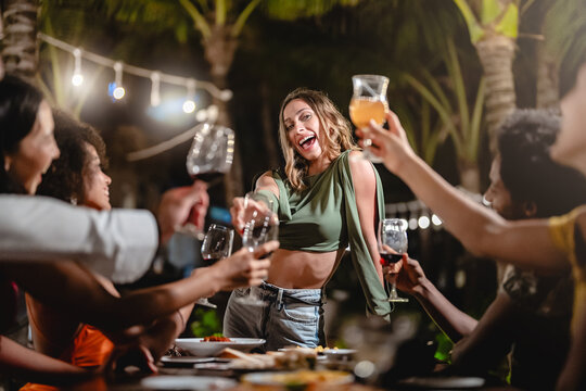 Group of diverse friends raising glasses in a joyful toast at a tropical outdoor party, celebrating together under palm trees and string lights