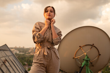 A fashionable woman in a beige outfit standing confidently on a rooftop during golden hour. The...