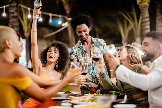 Group of diverse friends raising glasses in a joyful toast at a tropical outdoor party, celebrating together under palm trees and string lights