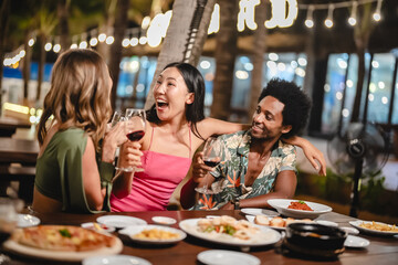 Group of happy friends sharing laughs and drinks at an outdoor dinner party under palm trees and string lights, enjoying food and festive vibes during a tropical night celebration