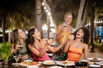 Group of joyful women toasting with wine glasses at an outdoor tropical party at night, smiling and celebrating together under string lights surrounded by palm trees and a festive atmosphere