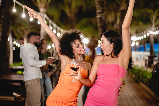 Two joyful women dancing with wine glasses during a fun tropical night party, surrounded by palm trees and string lights, celebrating friendship and enjoying the beach nightlife