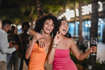 Two joyful women dancing with wine glasses during a fun tropical night party, surrounded by palm trees and string lights, celebrating friendship and enjoying the beach nightlife