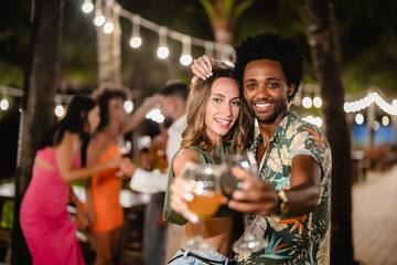 Smiling man and woman posing together while holding drinks at an outdoor night party, surrounded by friends dancing and celebrating under tropical palm trees and string lights