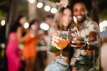 Smiling man and woman posing together while holding drinks at an outdoor night party, surrounded by friends dancing and celebrating under tropical palm trees and string lights