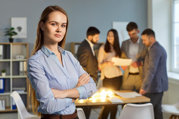 Portrait of confident young business woman looking at camera standing in office with people company employees working in background with glowing lightbulbs at table. Innovation and idea concept.