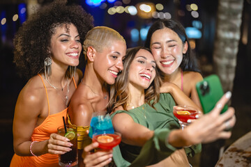 Four diverse young women enjoying a night out, smiling and taking a selfie together while holding colorful cocktails at an outdoor party.