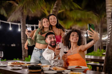 Group of friends taking a selfie while enjoying drinks and dinner at an outdoor tropical restaurant, smiling and celebrating under string lights.