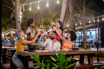Group of diverse friends raising glasses in a joyful toast at a tropical outdoor party, celebrating together under palm trees and string lights