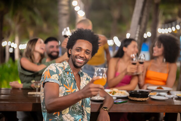 Smiling man raising a glass at an outdoor dinner party with friends, celebrating and enjoying food and drinks at night under string lights in a tropical setting