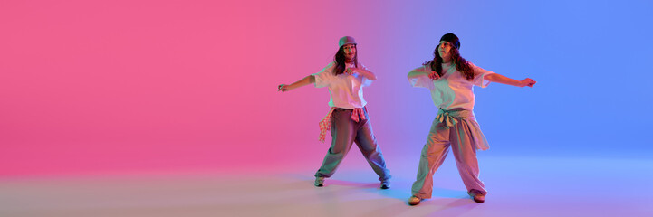 Two girls performing synchronized hip hop routine under bright neon lighting against gradient...