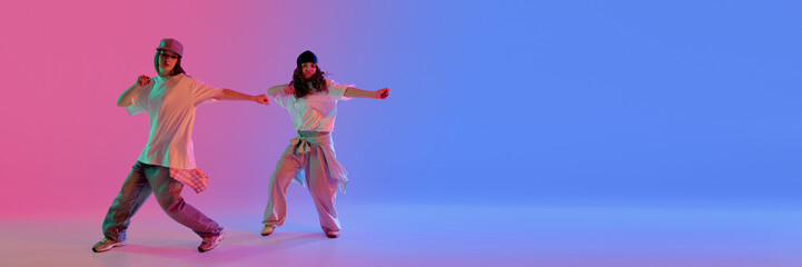 Confident young female dancers moving in sync under pink and blue lights against gradient...
