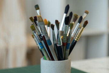 Watercolor, oil, and acrylic brushes collected in a single ceramic mug, bathed in workshop light