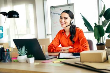 Plakat Smiling female entrepreneur working on laptop at her desk with packages in a bright modern office environment