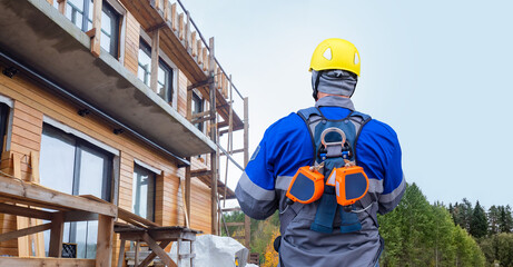 Construction worker in safety gear with fall protection equipment inspects wooden building...