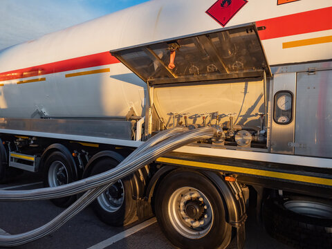 Fuel tanker truck with hazardous material sign, connected stainless steel hoses, control valves, and pressure gauges for liquid transfer – Logistics, Industrial Transport.