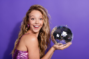 Young woman with blond hair and glowing top holding disco ball against purple backdrop