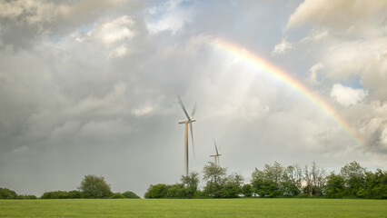 Side view of two wind turbines in the field under the cloudy sky with the rainbow