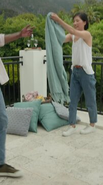 Vertical video: Couple entering frame grabbing teal blanket then shaking it open for terrace picnic