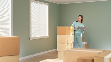 Woman is engaged in moving. Girl with boxes in empty room. Lady moved things to new apartment. Moving to purchased apartment. Relocation to new house. Female is engaged in relocation process