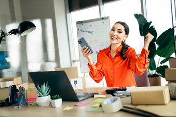 Successful female entrepreneur in modern office celebrating achievements with smartphone and...