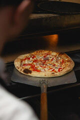 A close-up view of a pizza being removed from a stone oven using a classic wooden-handled peel. The perfectly puffed and charred crust, along with toppings like mushrooms, red bell peppers, and ham