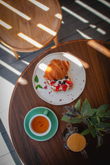 Overhead view of a fresh croissant with cream and berries, served with a cup of tea and a teapot on a sunny wooden table. Cozy breakfast mood in natural light