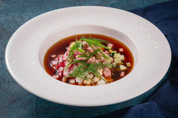A bowl of classic Russian okroshka soup made with kvass, ham, radish, cucumber, eggs and herbs. Served cold in a white bowl on a blue background