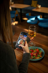 A young woman photographs her food with a smartphone while dining at a restaurant. A glass of white wine and a vibrant dish with vegetables are on the table