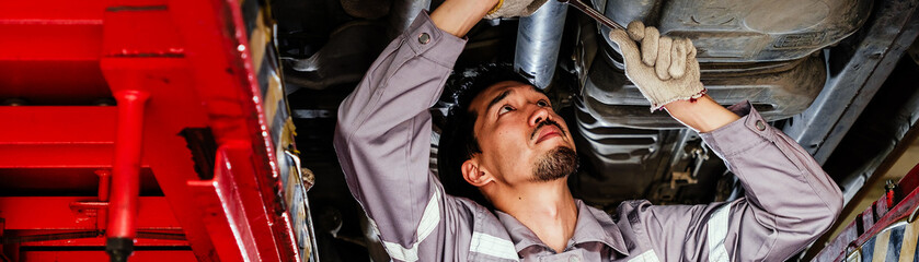 Auto mechanic in gray uniform tightening components under a raised car in a repair shop, performing hands-on maintenance with a wrench during routine vehicle inspection and mechanical servicing.