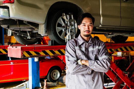 Confident auto mechanic in gray uniform stands with arms crossed in front of elevated vehicle at repair shop, representing expert service, professional inspection, and car maintenance confidence.