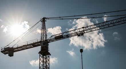 Tower crane silhouette against a bright sky with scattered clouds and sun flare