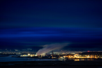 Fototapeta premium Industrial shot from the Cheshire Plains at night, featuring a brightly lit complex with steam rising against a deep blue sky, overlooking a body of water and distant city lights