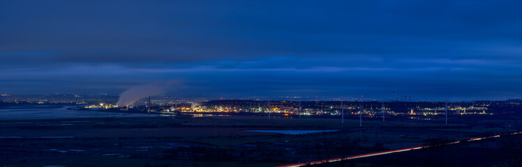 Industrial panorama from the Cheshire Plains at night, featuring a brightly lit complex with steam rising against a deep blue sky, overlooking a body of water and distant city lights